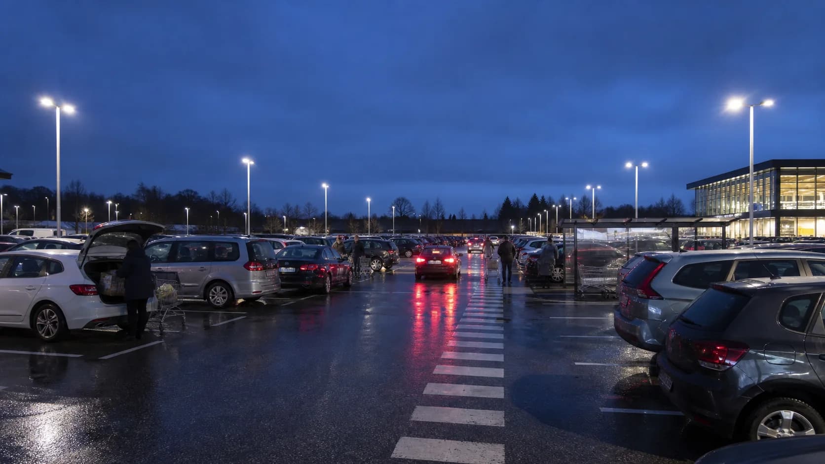 Occupied commercial car park at dusk with bays, pedestrian route, entrances and pole locations visible; avoid an empty generic lot or exaggerated brightness.