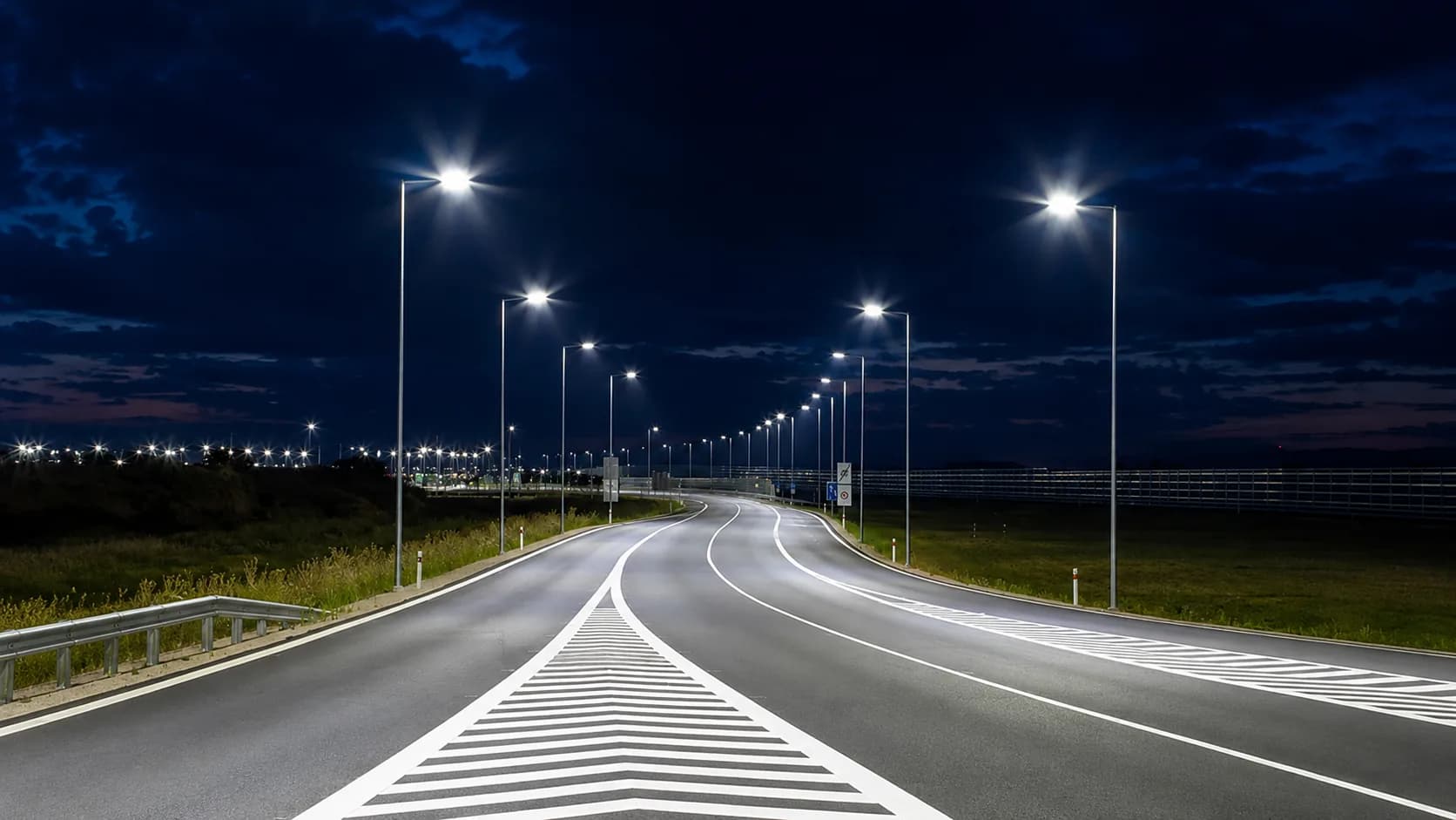 Street lighting poles illuminating a public road at dusk.