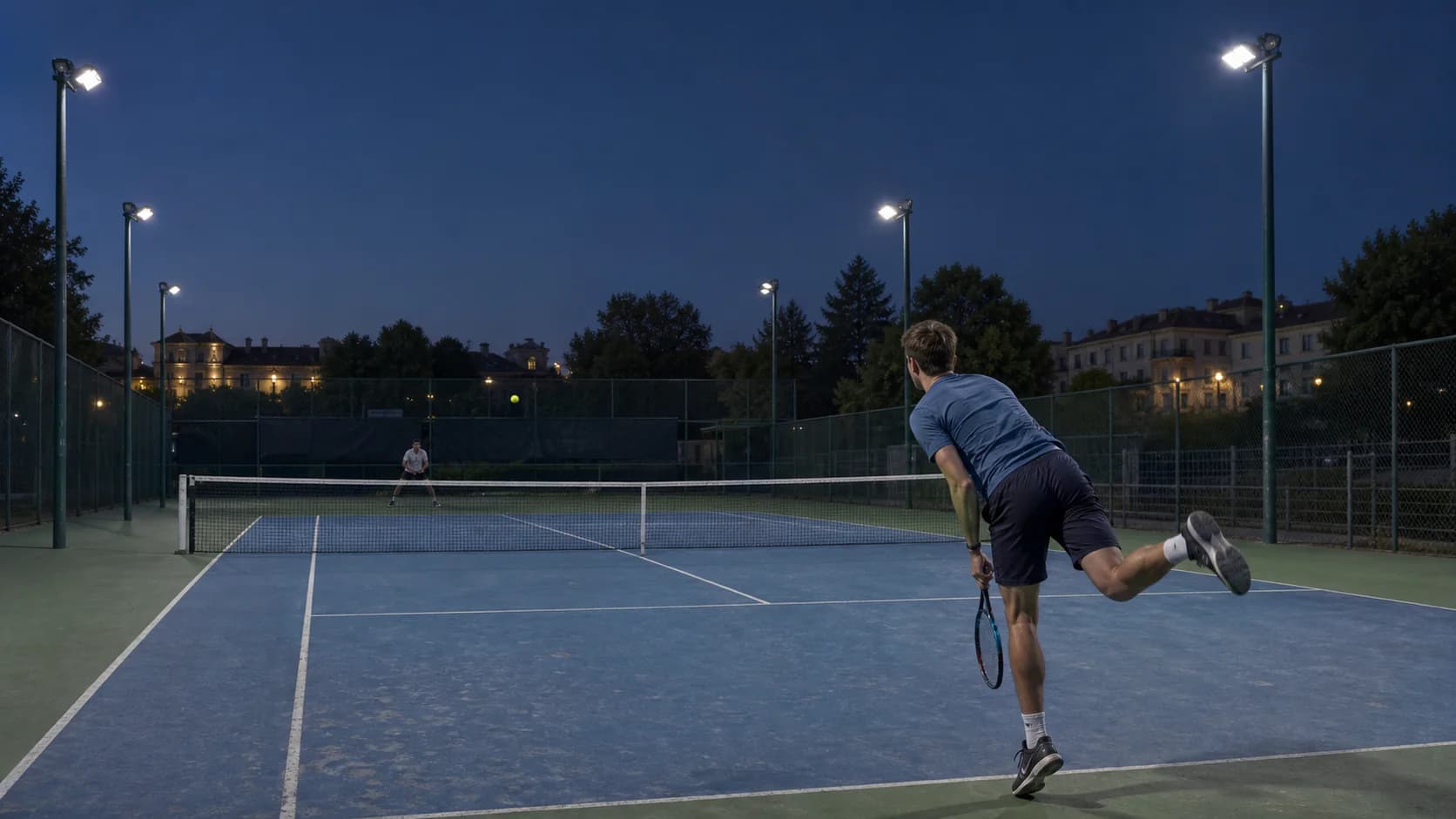 Real outdoor tennis courts at evening play, photographed across the net so side-pole positions, fences and adjacent courts are visible; no staged tournament crowd.